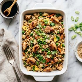 Cauliflower fried rice with chicken in a white ceramic dish on marble surface with green onions and sesame seeds