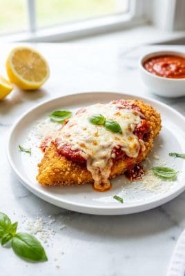 Single serving of oven baked chicken parmesan on a white plate with melted mozzarella and marinara sauce